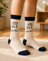 Pair of cream and black crew socks featuring a funny Arabic meme character holding a broken heart with the phrase "أصلاً عادي" on a white background.