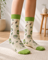 Pair of cream and green Matcha Tea Crew Socks featuring matcha cups, bamboo whisks, tea leaves, and matcha tin illustrations on a white background.